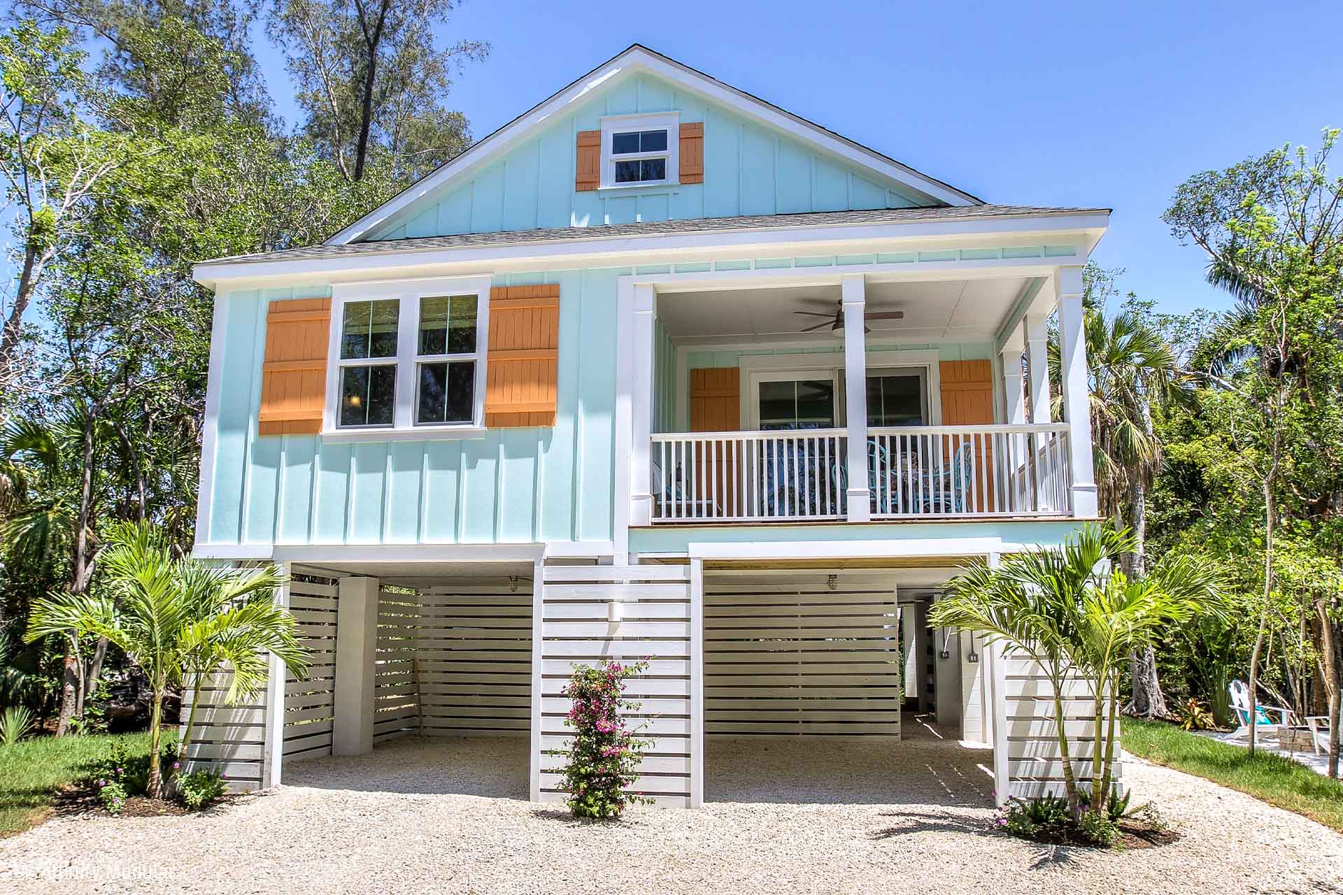 Light blue elevated modular home with orange shutters, white railings, and palm trees in a sunny, tropical setting.